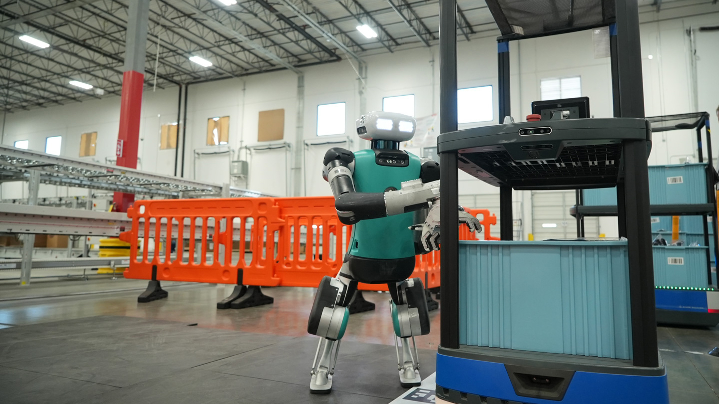 Agility Robotics Digit humanoid moving a tote beside an autonomous mobile robot in a warehouse workcell